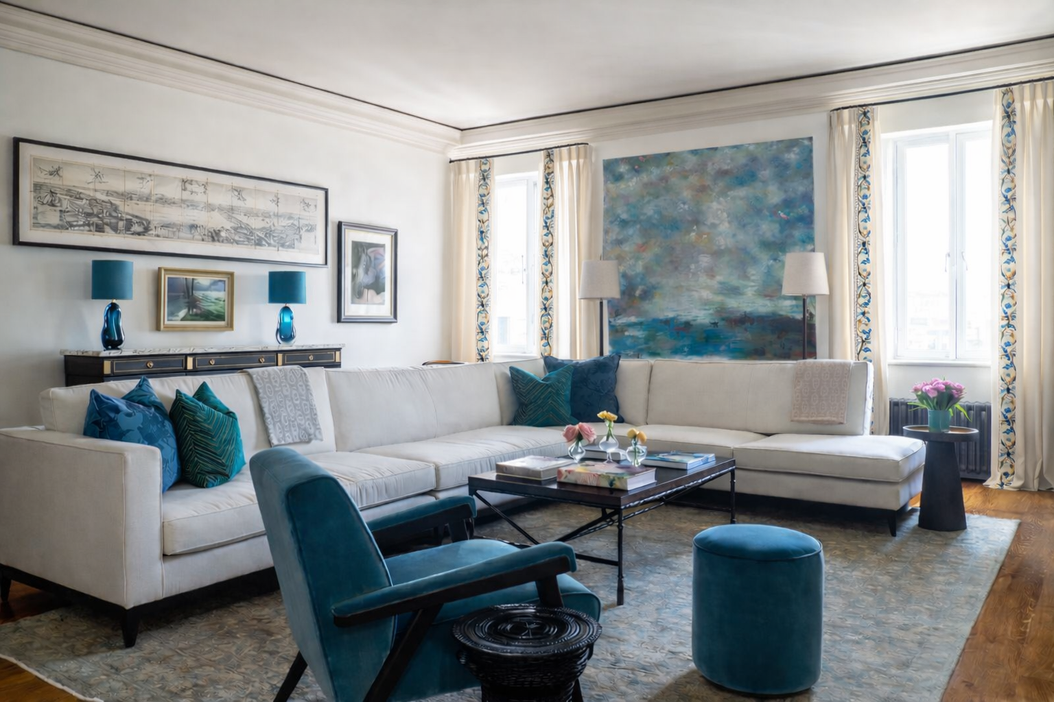 Sophisticated living room with cream curtains featuring embroidered botanical borders, cream sofas, teal velvet accents, and large abstract blue painting