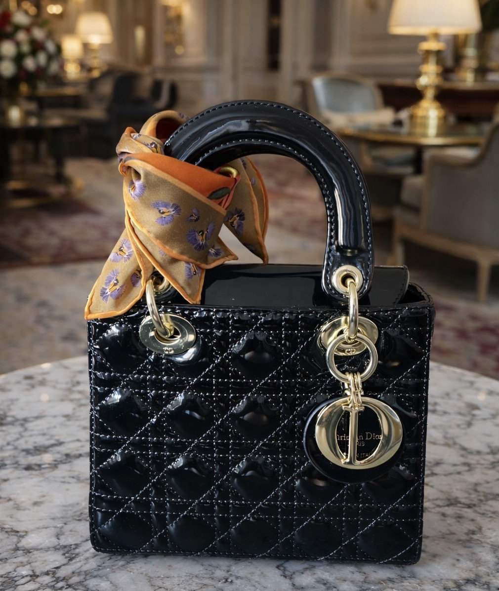 Gold Bird In Flight silk scarf with lavender flower and bee pattern tied around black patent Lady Dior handbag handle with gold hardware in elegant hotel interior