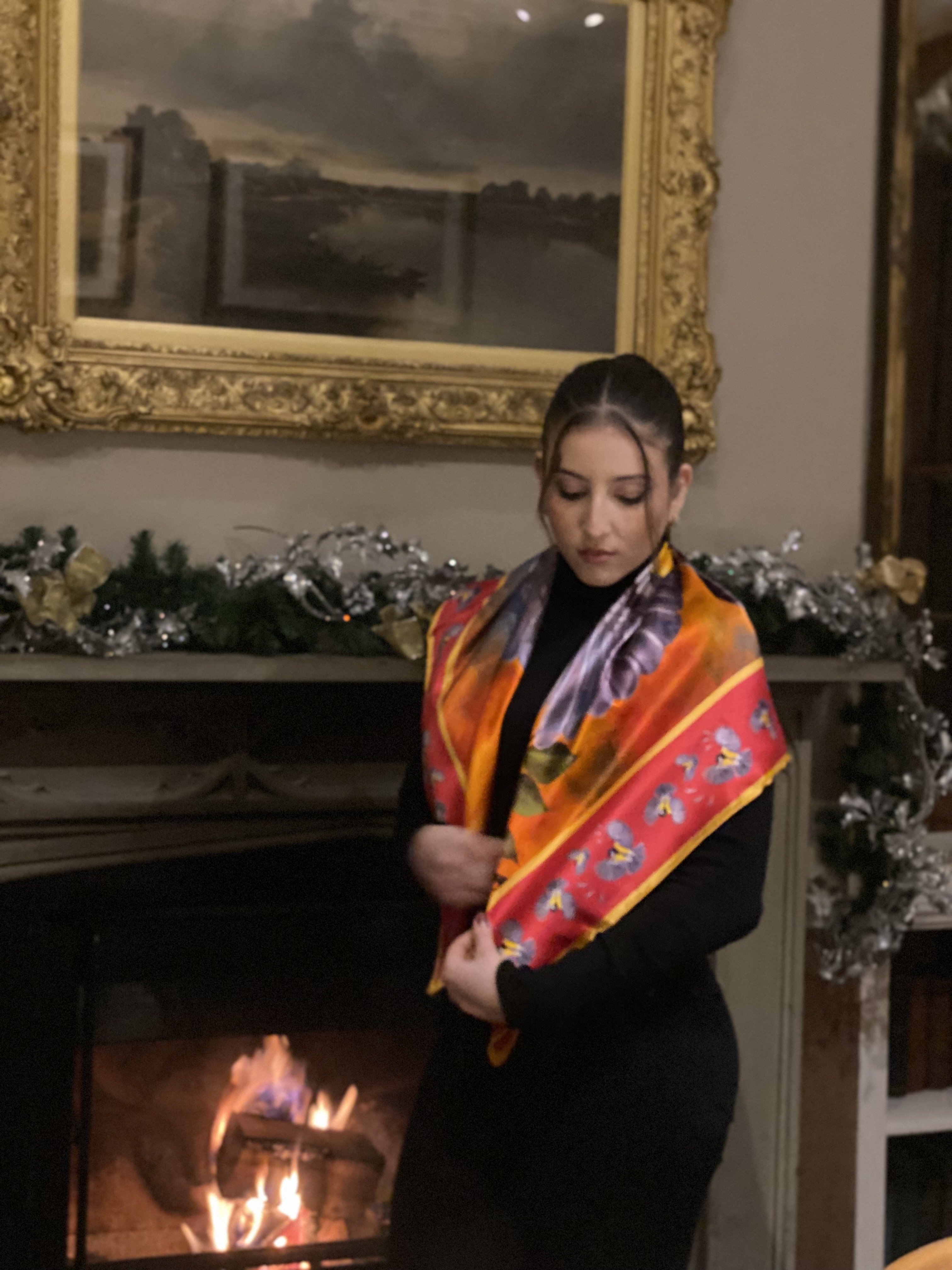 Woman wearing Bird In Flight silk scarf in red colourway draped over shoulders with black turtleneck by fireplace in elegant interior with gilt-framed painting