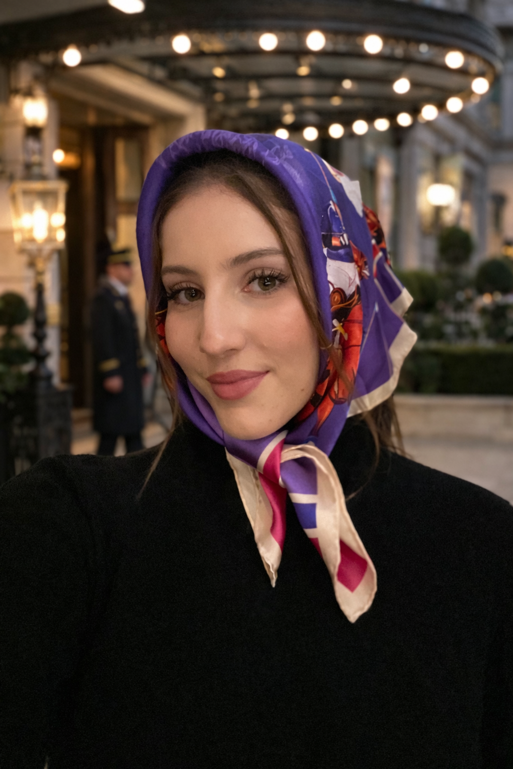 Woman wearing purple Equestrian Elegance silk scarf as a headscarf tied under the chin with black coat outside luxury hotel